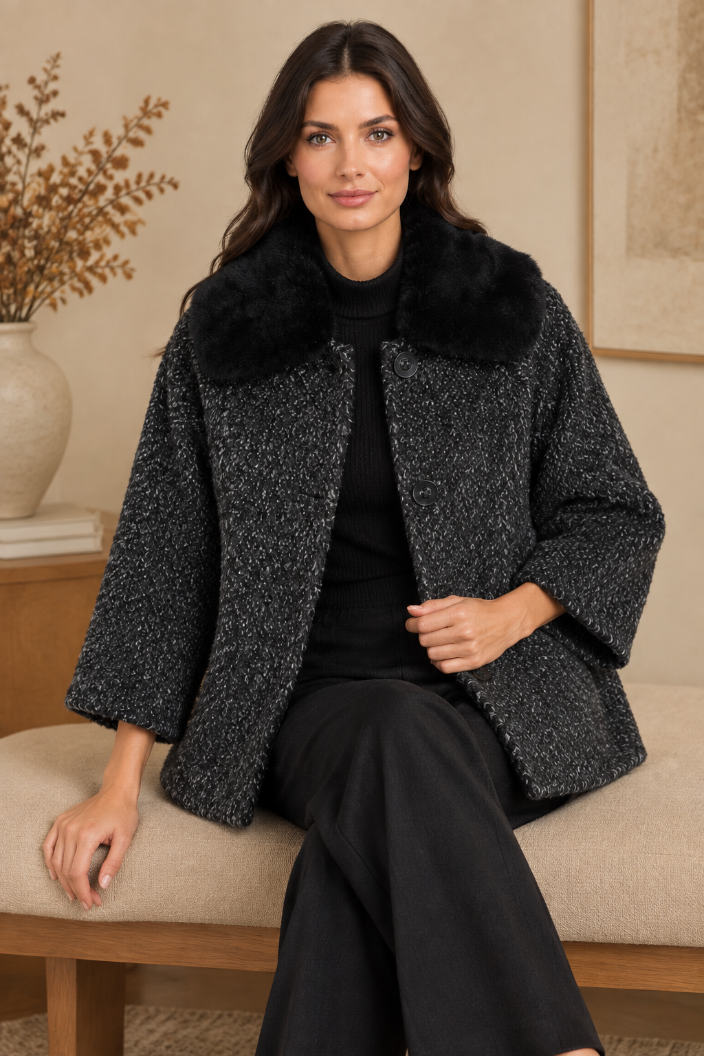 Model sitzt in einem dunkelgrauen Bouclé-Wintermantel mit schwarzem Fellkragen